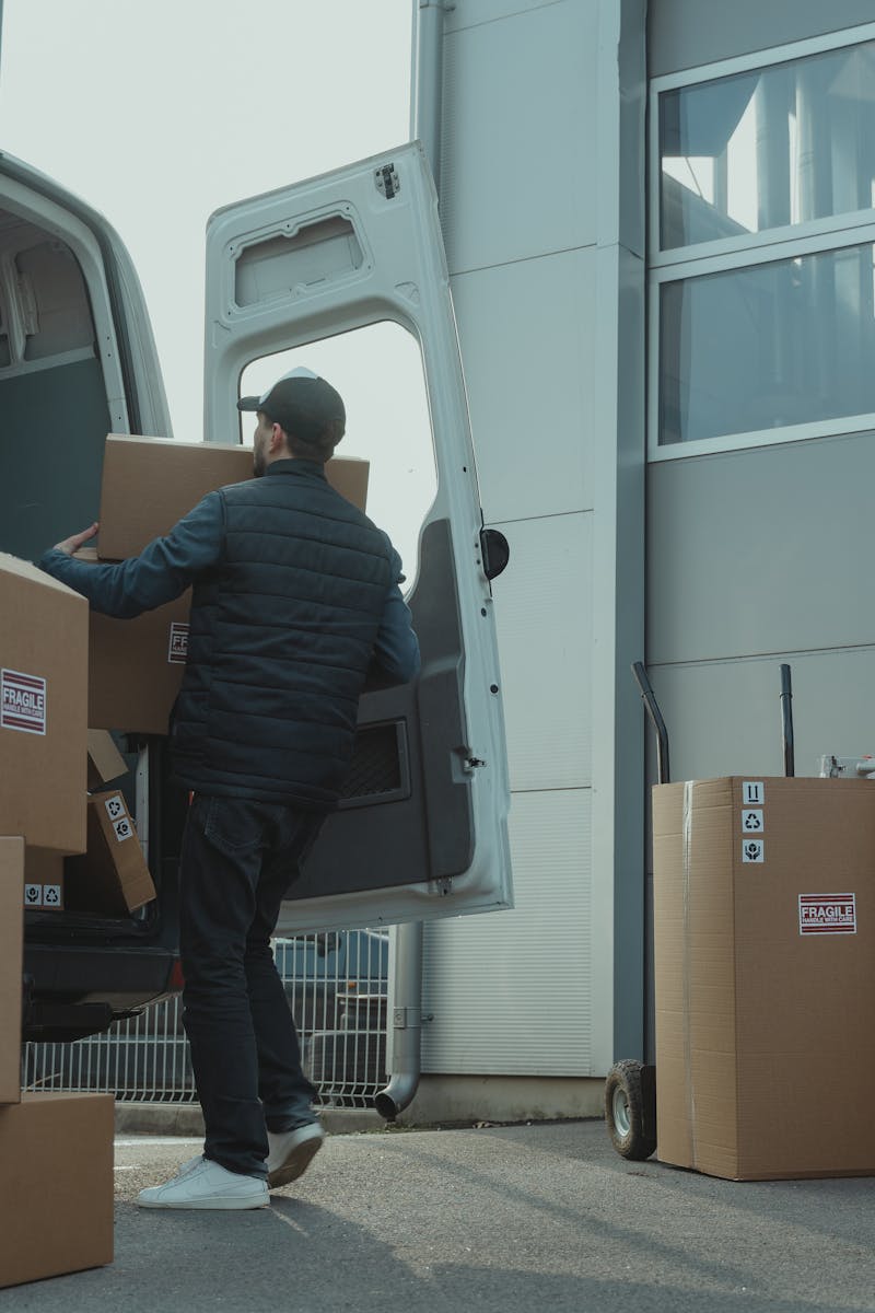 A delivery man unloading cardboard boxes from a van at a warehouse during the day.